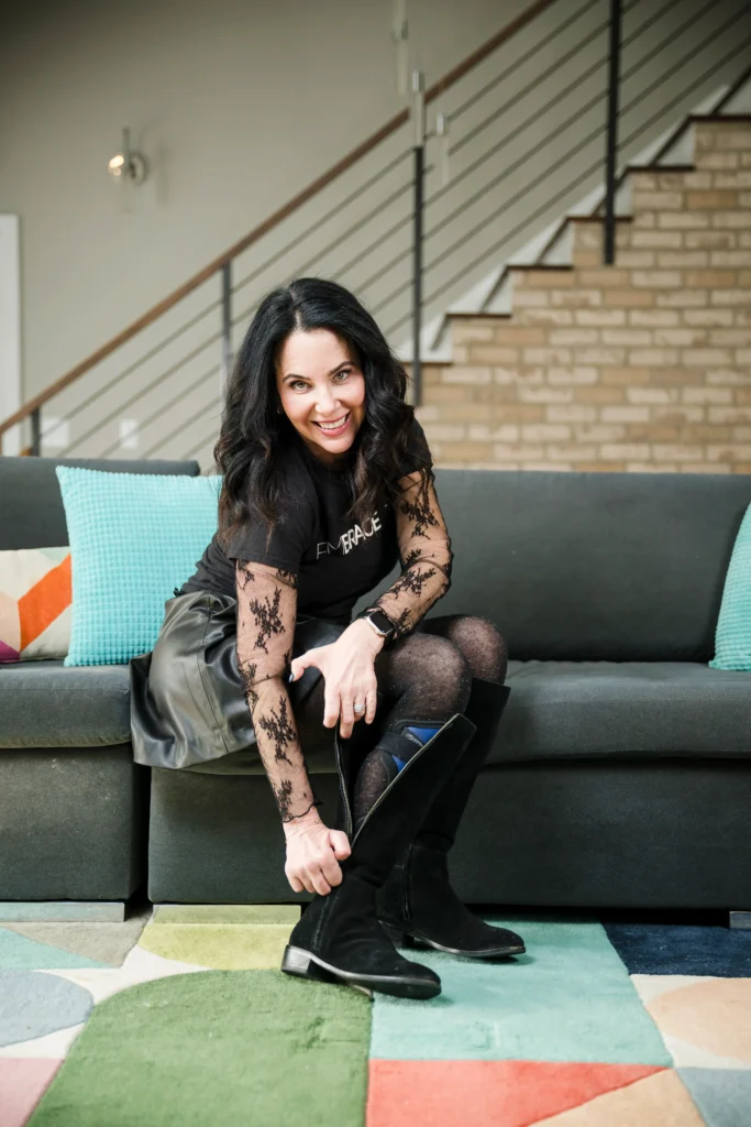 Lainie Ishbia sitting on a grey sofa and zipping up a black knee-high boot, wearing a black graphic tee that says "Embrace" with lace sleeves, a leather skirt, and patterned tights.