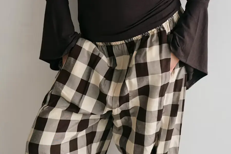 Close-up of a person's lower torso wearing a black long-sleeve top and checkered brown, cream, and gray plaid trousers with hands tucked in the pockets