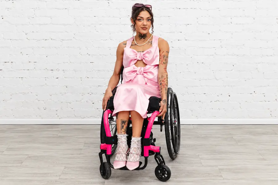 A woman with dark curly hair pinned up, sunglasses resting on her head, and a pearl necklace sits facing the camera in a lightweight black wheelchair with bright pink frame details. She wears a sleeveless, knee-length pink satin dress with two oversized bows stacked down the front and a small cutout between them. Her pink pointed-toe heels are decorated with pearl ankle straps and worn over white lace socks. Tattoos are visible on her neck, chest, and arms, including a butterfly at the base of her throat and red floral designs on her forearm. She stands out against a white-painted brick wall and a pale wood-look floor.
