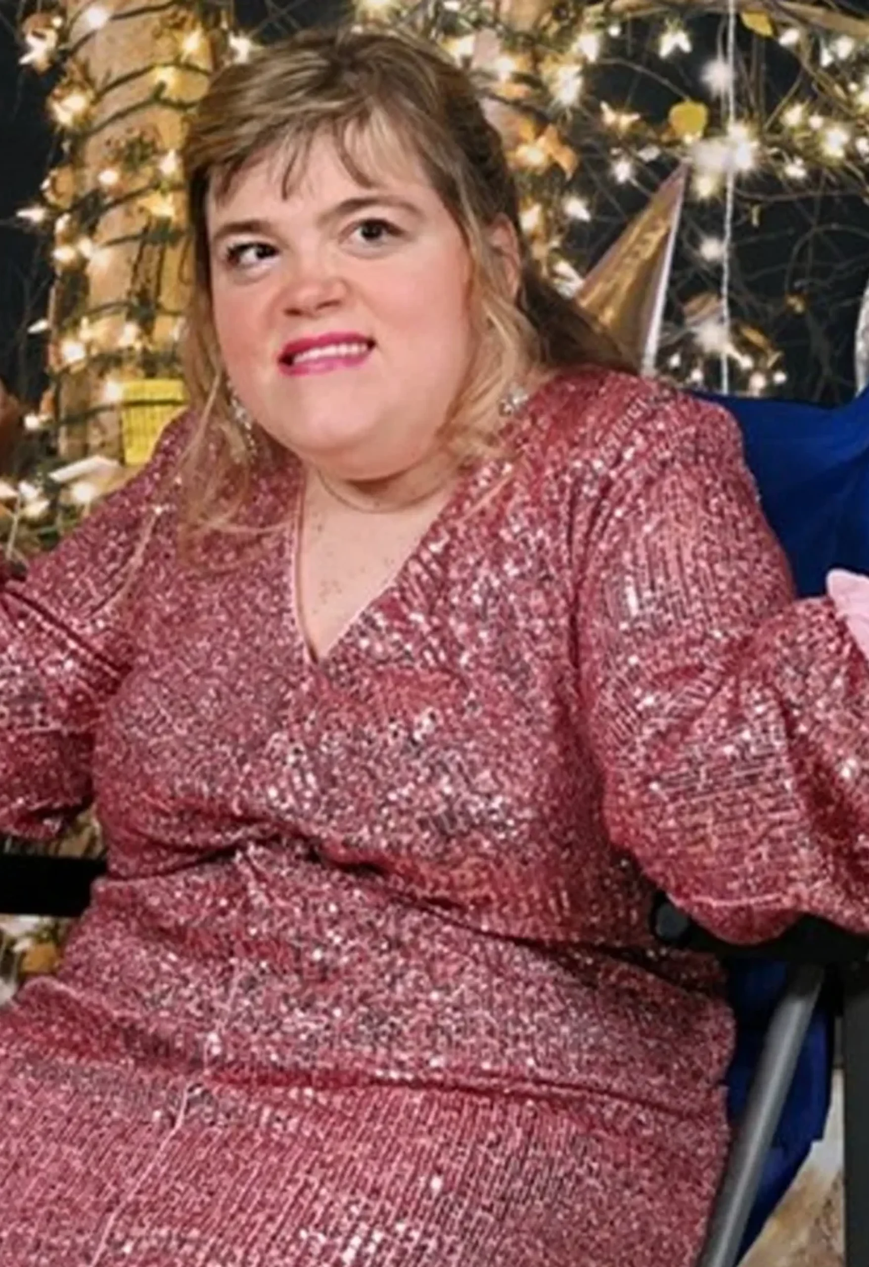 A joyful woman in a wheelchair posing in a sparkling rose gold sequin adaptive evening gown surrounded by glowing holiday lights and glass ornaments.