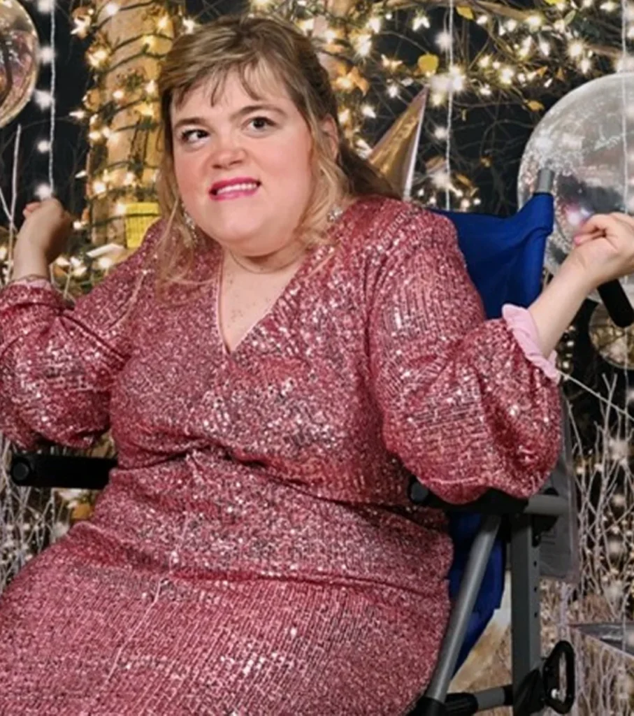 A joyful woman sitting in a blue chair, wearing a glittering pink sequin dress with long sleeves, smiling in front of a festive backdrop of sparkling fairy lights and holiday decorations.