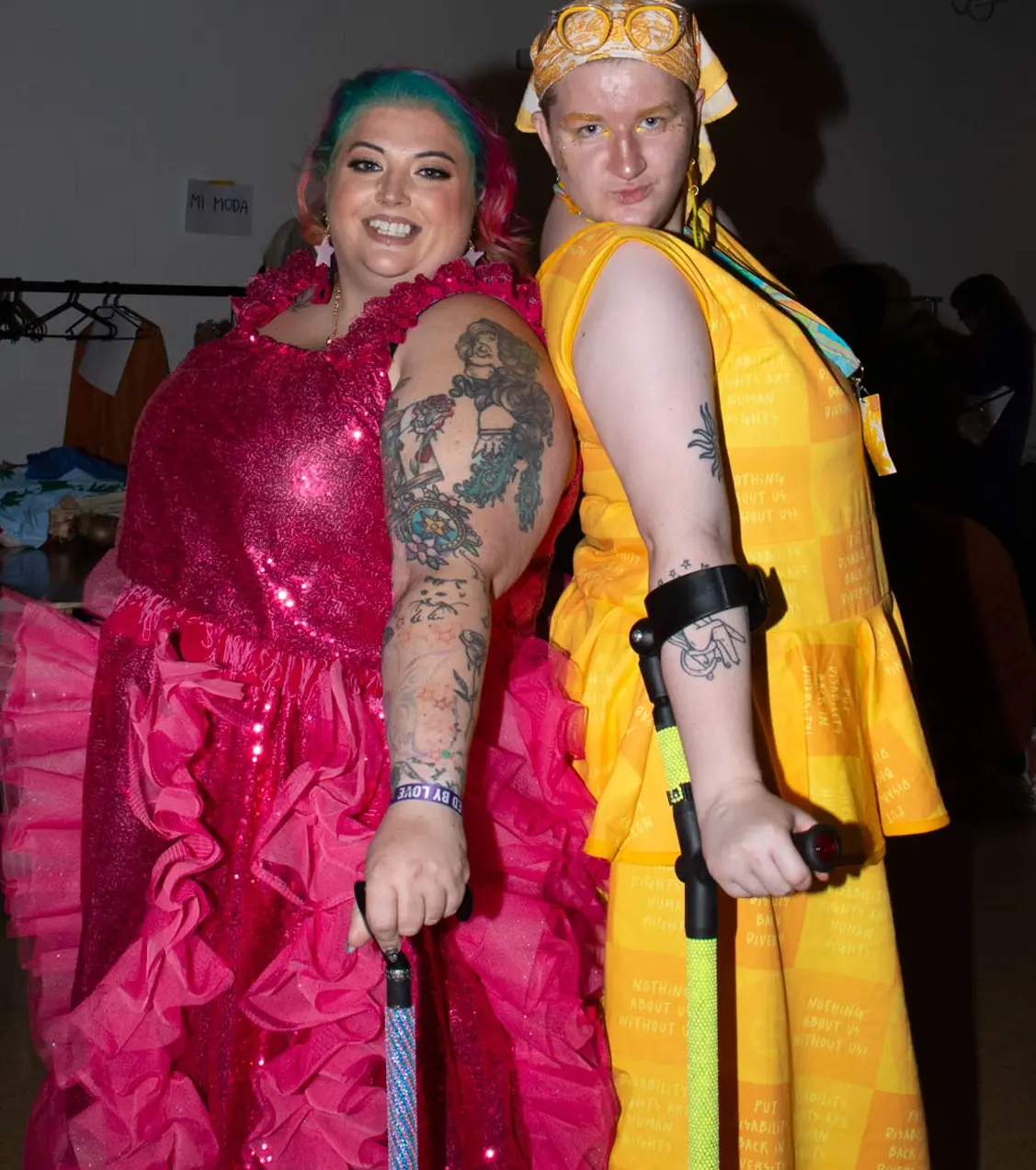 Two brightly dressed individuals posing back-to-back at a fashion event; one wears a shimmering hot pink ruffled dress and holds a cane, while the other wears a yellow patterned dress with matching headscarf and uses forearm crutches.