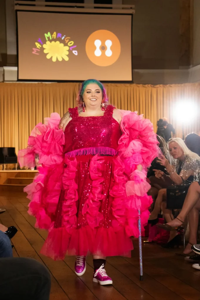 A plus-size model walking the runway with a sparkling cane, wearing a shimmering hot pink sequin dress with voluminous ruffled tulle sleeves at the May Marigold fashion show.
