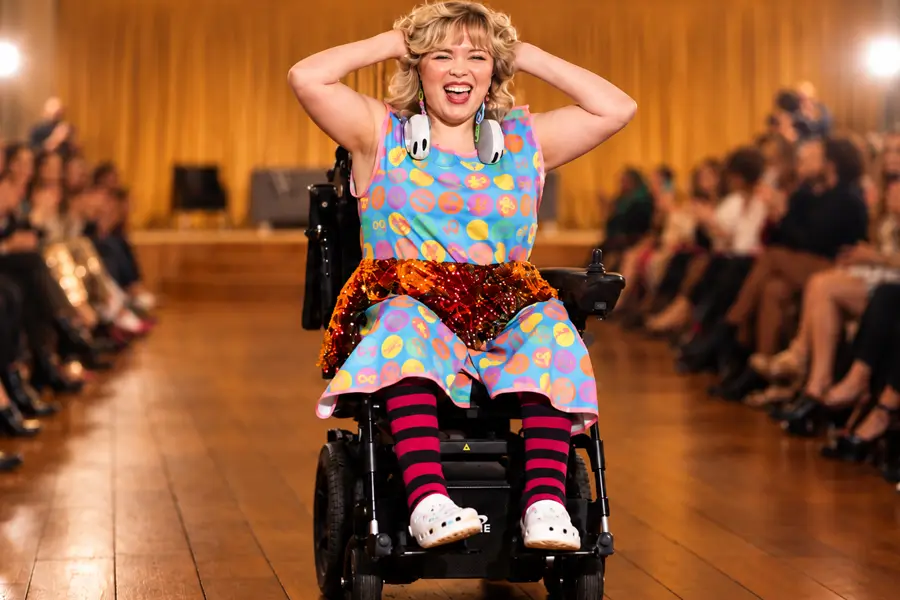 At a fashion show in a warmly lit hall with wooden floors, a woman with voluminous, curly blonde hair and bangs rides toward the camera in a black power wheelchair, laughing with both hands pressed to her head. She wears a sleeveless blue dress with pink trim, printed with rainbow-colored circles containing small symbols, and a band of orange and red sequins across the hips. White headphones hang around her neck, and she completes the look with red-and-navy striped knee socks and white clogs. Behind her, a projector screen displays the ‘May Marigold’ logo with a yellow flower beside an orange circular emblem. Audience members sit along the right side of the runway, and a photographer and a bright stage light are visible in the background