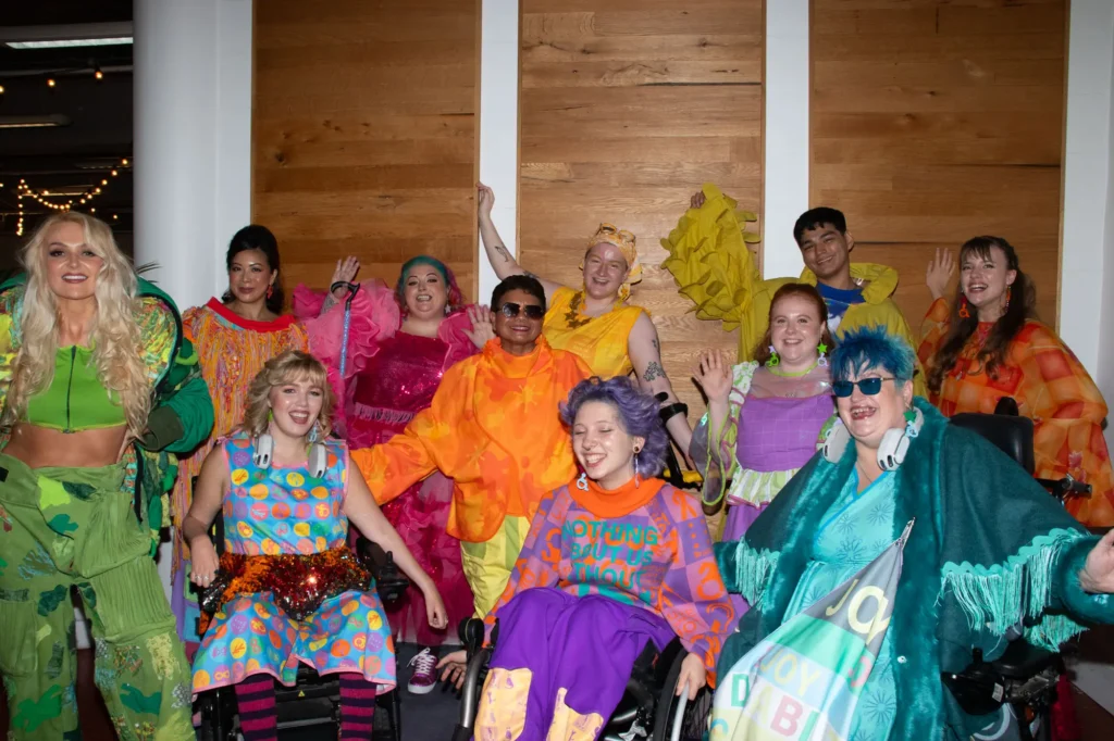 A diverse group of models posing together backstage in eclectic, bright streetwear and adaptive clothing designs against a wooden wall panel backdrop.