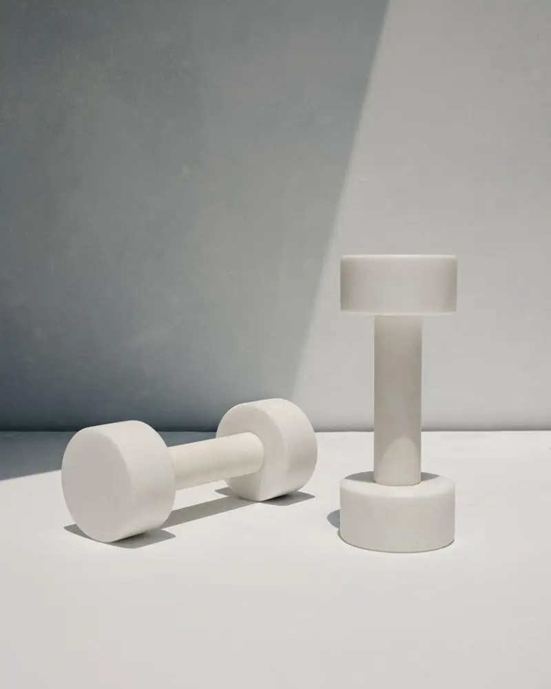 Two minimalist cream-white stone dumbbells, one lying on its side and one standing upright, on a pale surface against a gray wall split by a diagonal band of light and shadow.