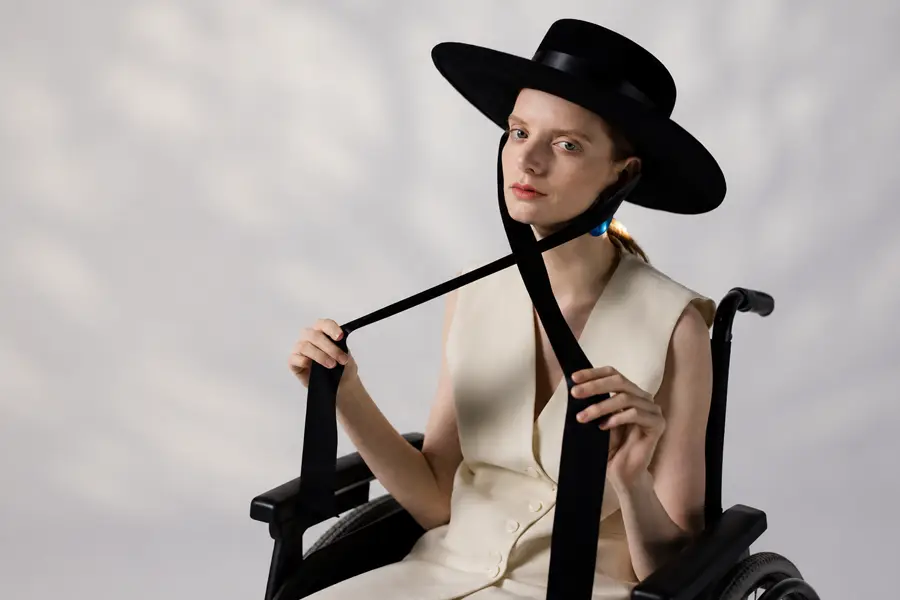 A model wearing a wide-brimmed black hat with a long strap and a cream sleeveless outfit poses in a wheelchair against a neutral studio backdrop.