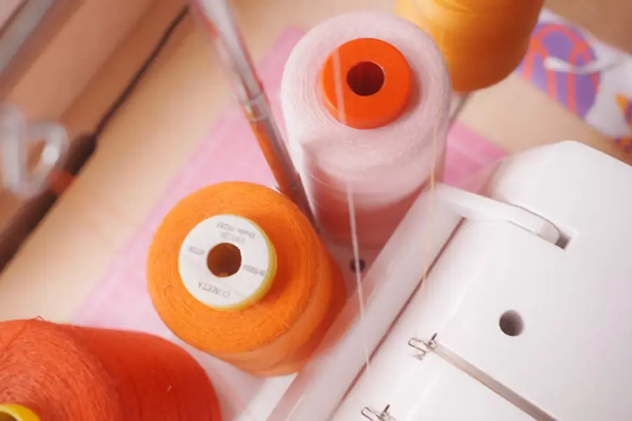 Close-up of large spools of orange, pale pink, and yellow-orange thread threaded onto a white sewing machine, on a pink cutting mat.