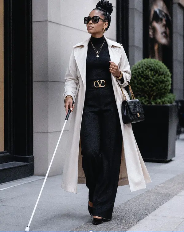 A chic, blind Black woman walking confidently down a city sidewalk while navigating with a long white cane. She is elegantly dressed in a black turtleneck jumpsuit with a gold logo belt, a long cream-colored trench coat, large dark sunglasses, and gold jewelry, while carrying a black quilted handbag.