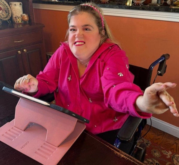 A blonde woman with a disability wearing a vibrant pink adaptive button-up shirt while sitting in a wheelchair and using a tablet on a pink desk stand.