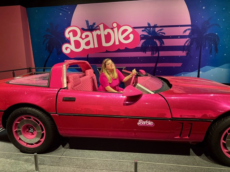 A smiling woman sitting inside a life-sized hot pink convertible Barbie car display under a large neon Barbie logo background.