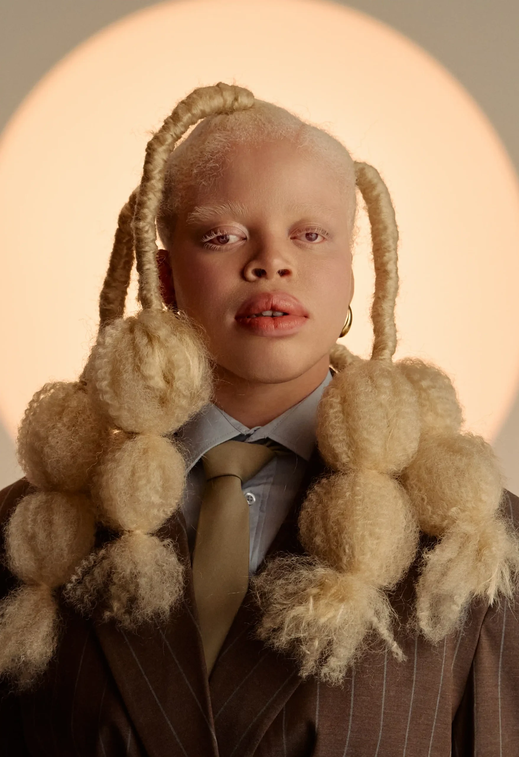 Close-up portrait of a Black model with albinism and styled blonde bubble-braid hair wearing a brown pinstripe suit jacket, light blue collared shirt, and olive tie.