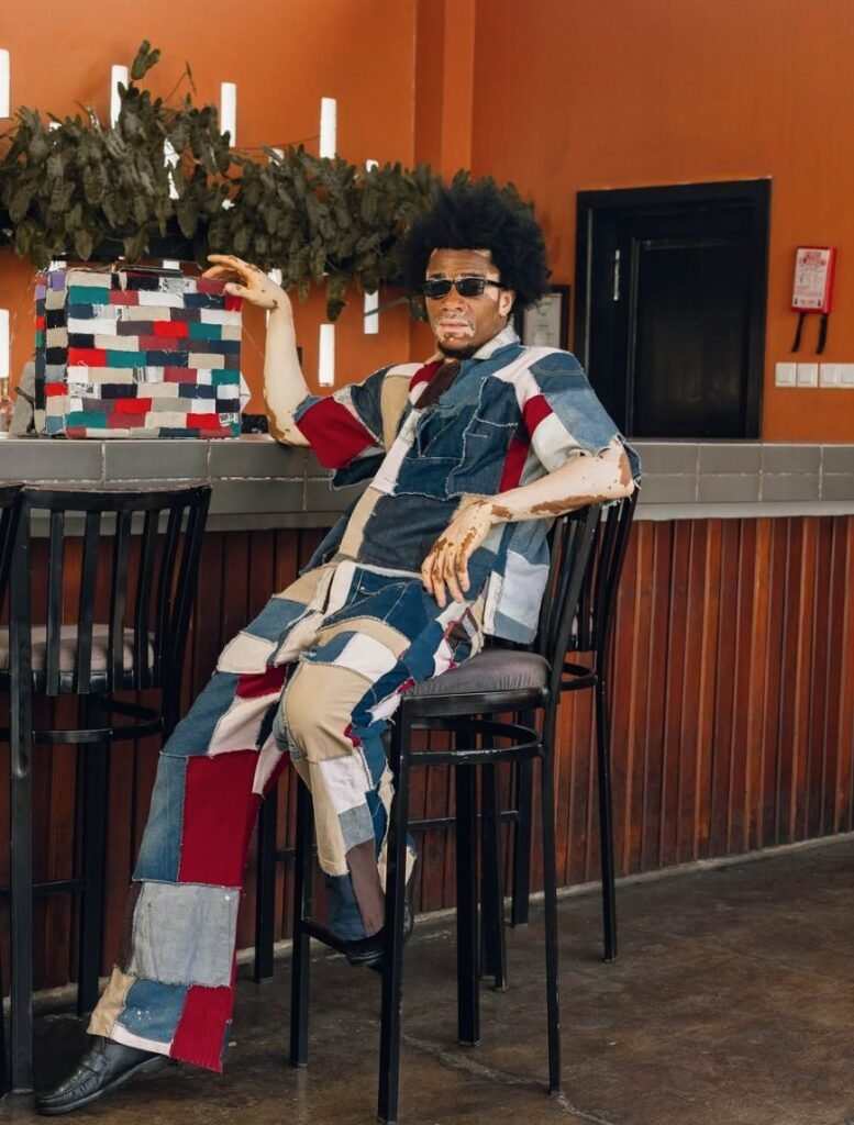 Person with a rounded afro and dark sunglasses lounges on a black bar stool, wearing a patchwork denim shirt and pants in blue, red, and tan, next to a matching patchwork bag on a bar counter against an orange wall.