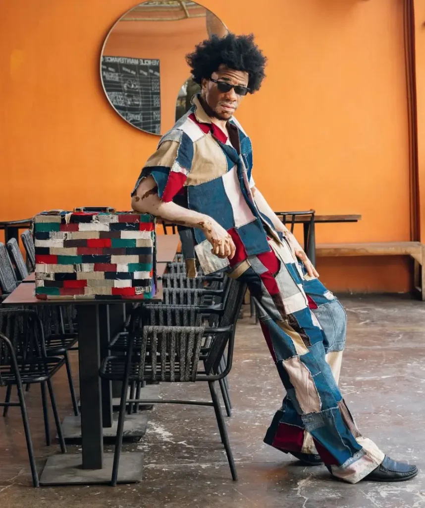 Full-length shot of fashion model Robert Essel wearing a colorful patchwork denim shirt and pants, leaning against a table next to a matching patchwork suitcase in a cafe.
