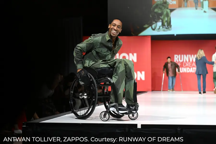 Antwan Tolliver of Zappos smiles broadly as he rolls down a white runway in a black wheelchair, wearing a matching olive-green jacket and pants, at a Runway of Dreams fashion show.