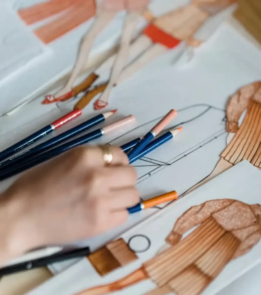 A hand with a gold ring reaches for blue colored pencils scattered across fashion design sketches in warm peach, orange, and red tones.
