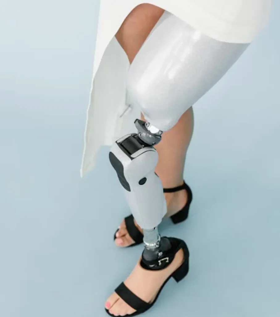 Cropped view of a person's legs in a white skirt and black block-heel sandals, with a sleek white and silver prosthetic leg with a visible knee joint, against a pale blue background.