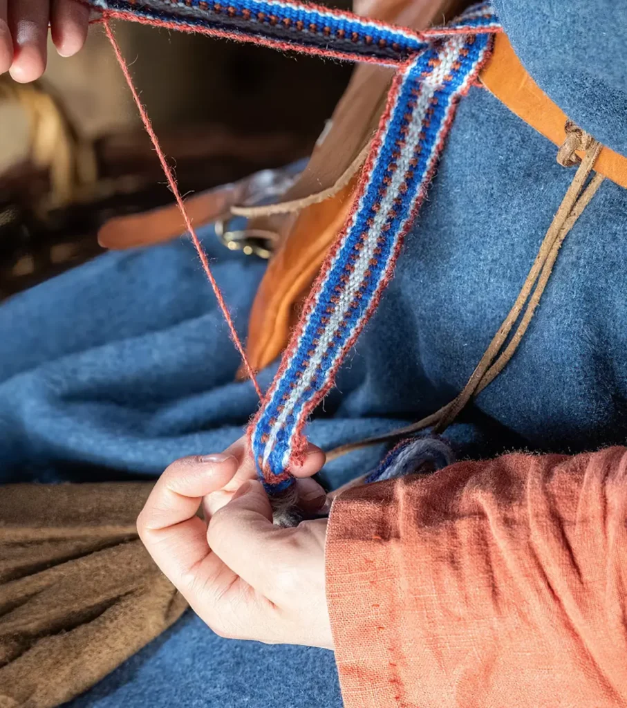 Close-up of hands working a narrow handwoven band in blue, white, and rust-red stripes, with a red thread pulled taut, against blue wool and an orange linen sleeve.