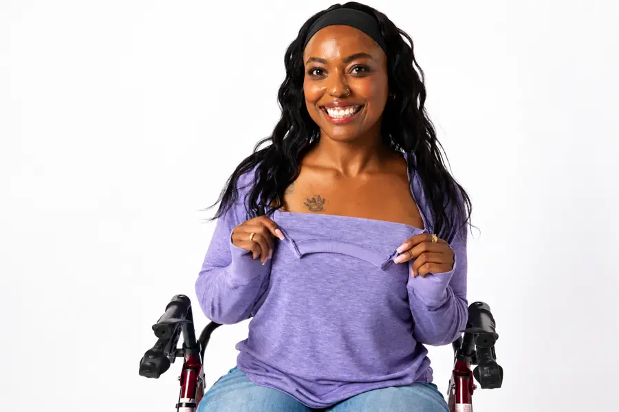 A woman with long, dark wavy hair and a black headband smiles warmly at the camera against a plain white studio background. She wears a soft heathered-lavender long-sleeve top with a wide neckline, and holds up a zippered opening at the front with both hands, showing how the garment opens. She has a small nose stud, gold rings, and a paw-print tattoo on her chest. Dark handles from a mobility device, likely a rollator or walker with red frame accents, are visible on either side of her, and she wears light blue jeans