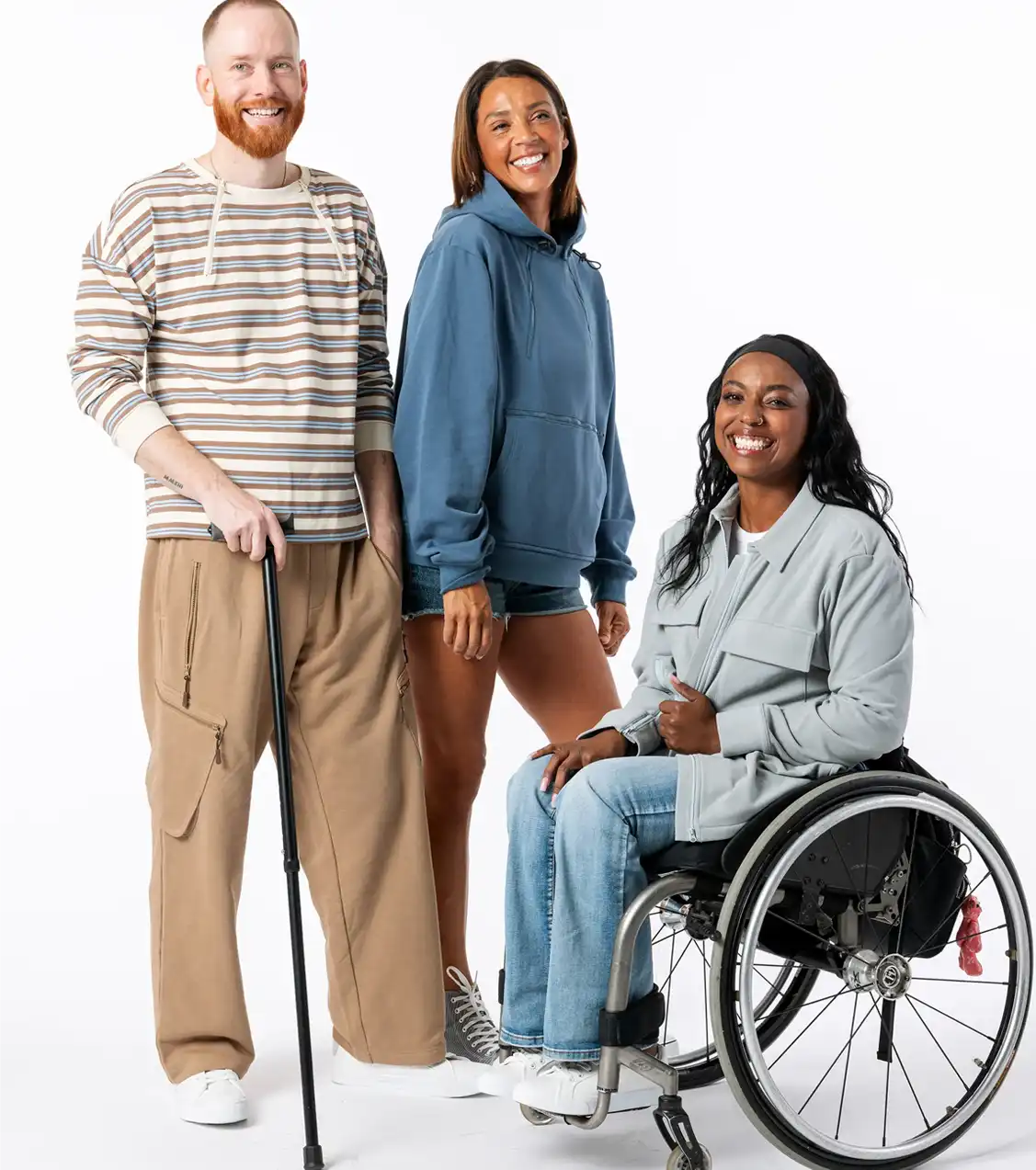 Three smiling individuals standing together on a white background, including a man using a walking cane, a woman in a blue hoodie, and a woman in a wheelchair wearing a grey jacket and jeans.