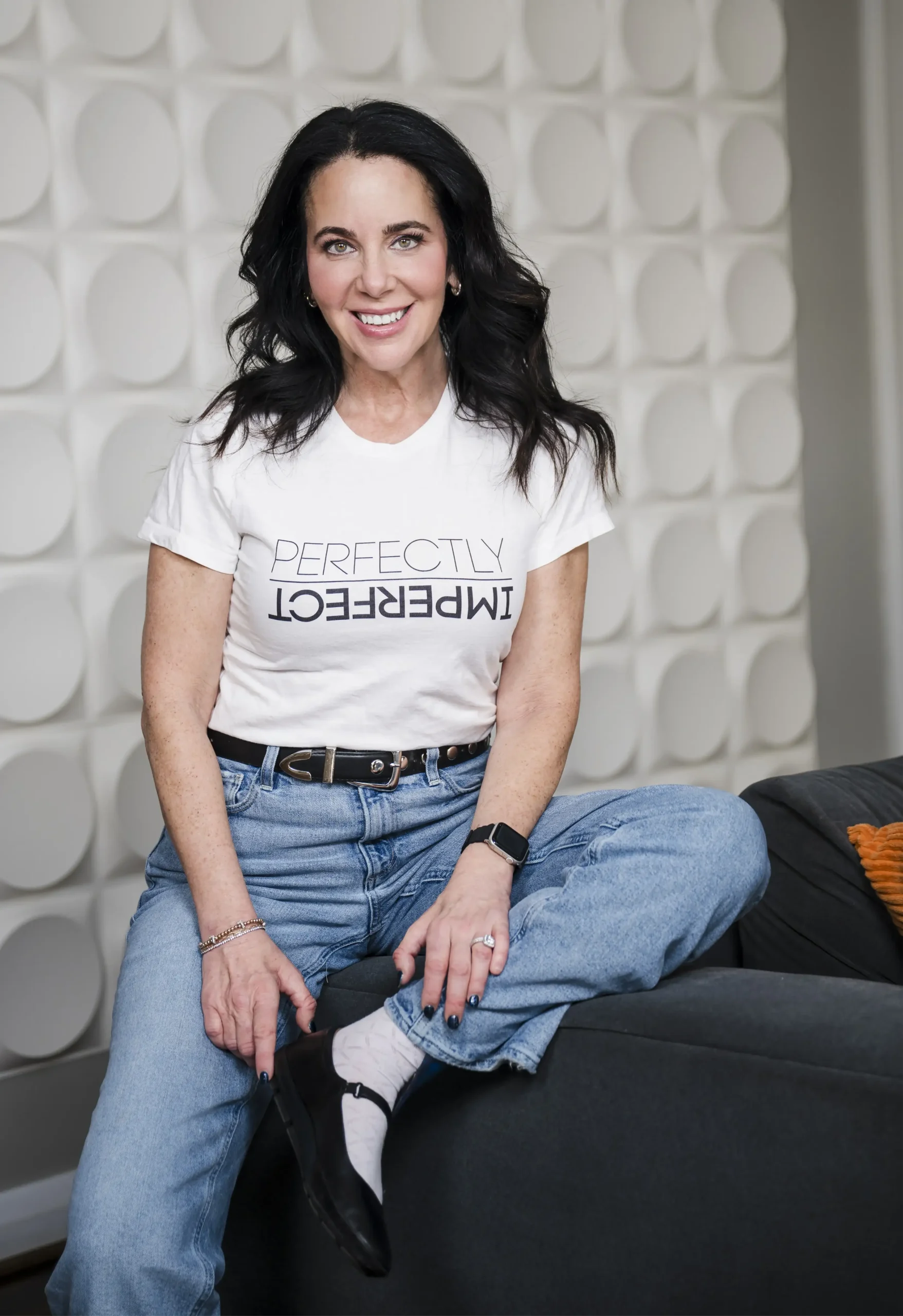Portrait of Lainie Ishbia sitting casually on the arm of a dark grey couch, wearing blue jeans and a white t-shirt with "Perfectly Imperfect" printed on it against a white textured wall.