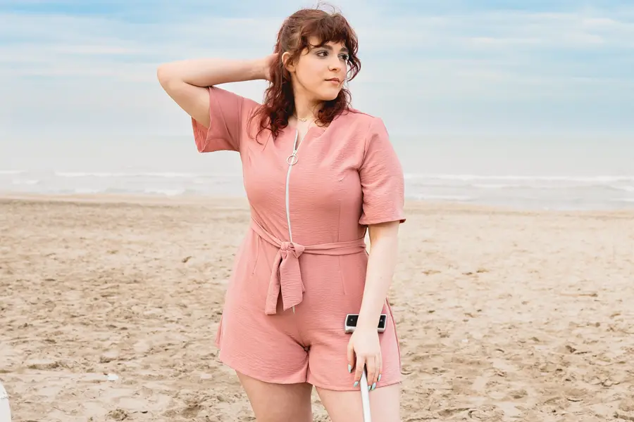 A woman with wavy, shoulder-length auburn hair and bangs stands on a sandy beach under a pale blue, cloudy sky, with gentle waves rolling in behind her. She wears a short-sleeved, dusty-rose romper with a zip front and a ring pull, and a fabric belt tied in a bow at the waist. One hand rests behind her head while the other holds a white cane planted in the sand, with a small device clipped to its handle. She has teal-painted nails and a delicate gold necklace, and gazes off to the side with a calm expression.