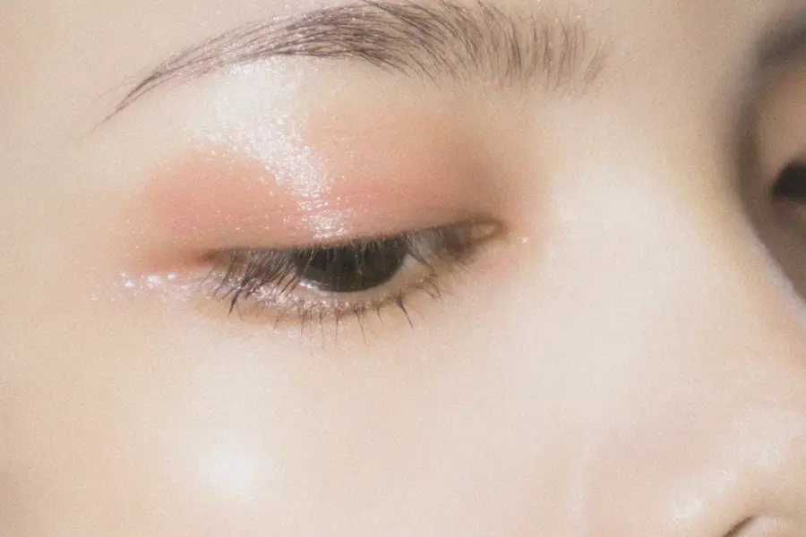 Extreme close-up of a person's half-closed eye with soft peach eyeshadow and a shimmery, glossy highlight on the lid, brow, and cheek.
