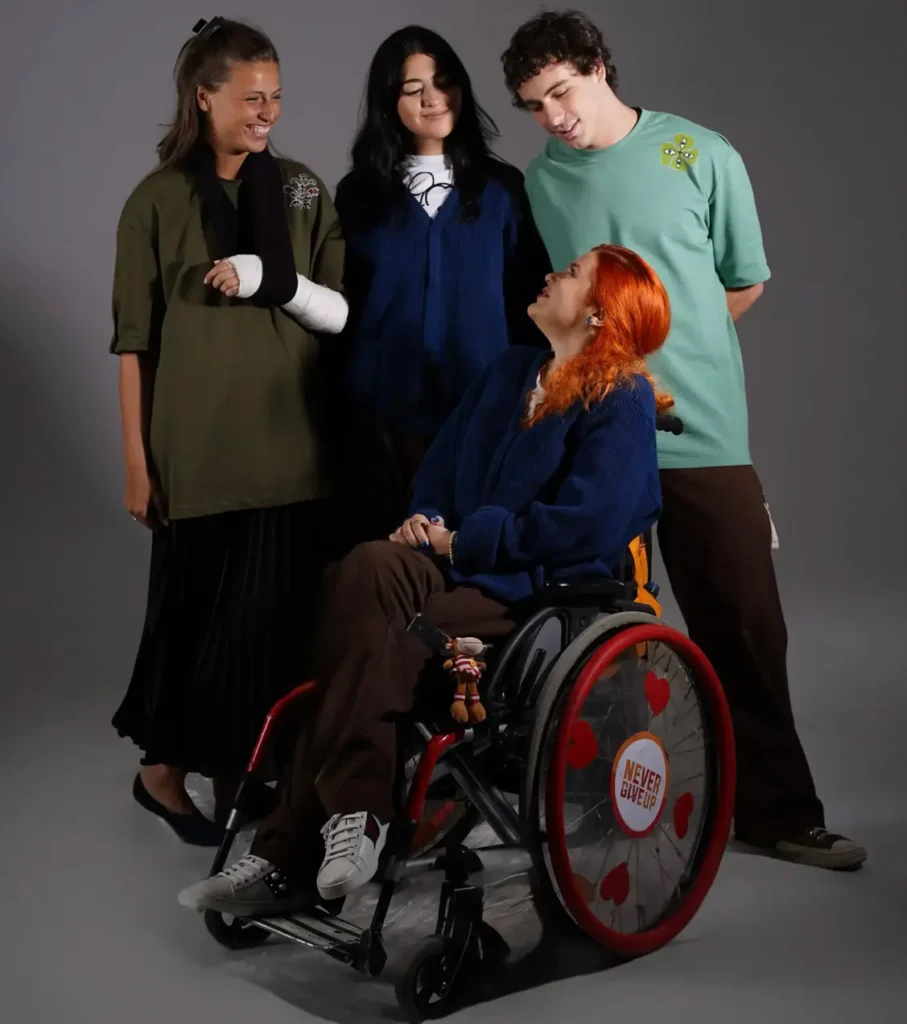 Four young adults pose together against a grey studio backdrop. A woman with bright red hair sits in a wheelchair looking up at three friends standing around her, including a woman wearing an arm sling over an olive green top, and a man in a mint green t-shirt.