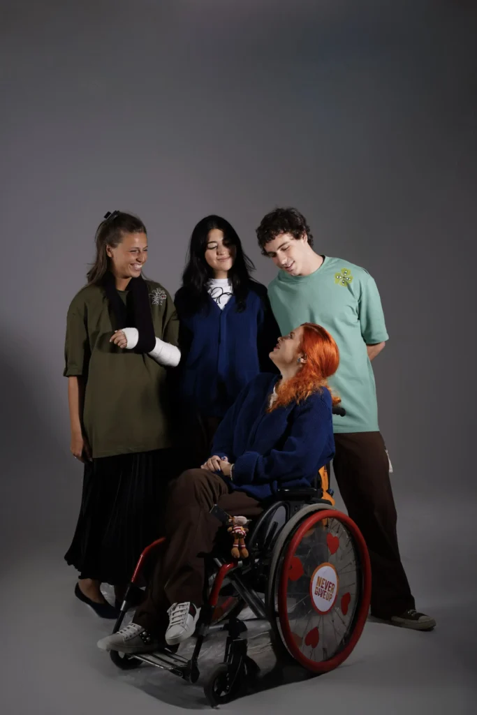 Four young adults pose together against a grey studio backdrop. A woman with bright red hair sits in a wheelchair looking up at three friends standing around her, including a woman wearing an arm sling over an olive green top, and a man in a mint green t-shirt.