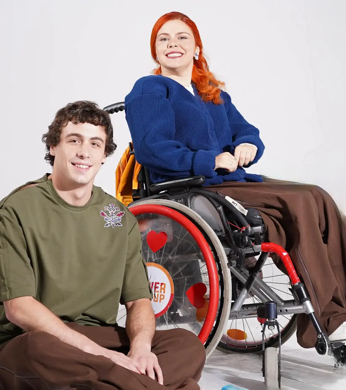 A smiling young man sits cross-legged on the floor wearing an olive green t-shirt and brown pants, next to a smiling woman with bright red hair using a wheelchair. She is wearing a cozy blue knit cardigan and brown wide-leg trousers.