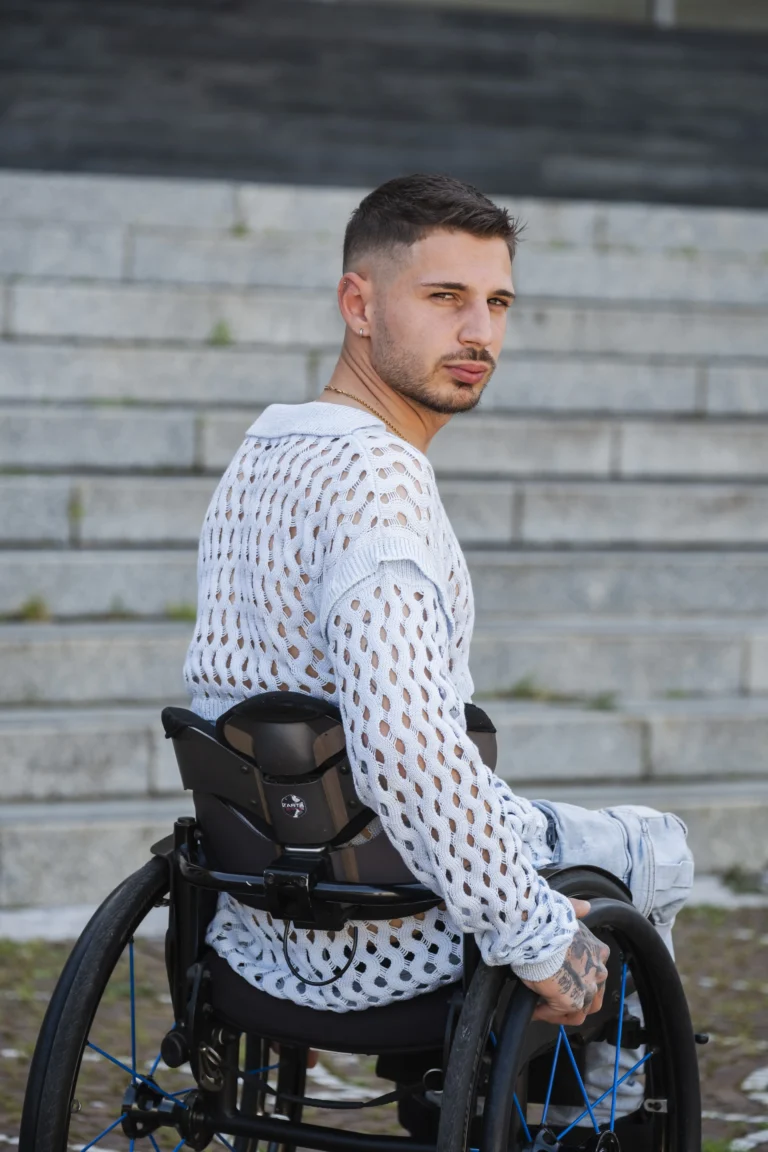 Over-the-shoulder portrait of a stylish young man in a wheelchair posing on stone steps, wearing an open-knit light blue crochet sweater and a gold chain necklace.