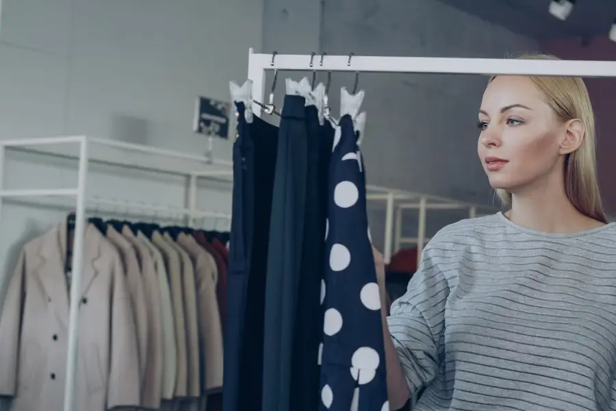 A woman browses clothing racks in a store, looking past hanging pants and a polka-dot skirt toward another rack of coats
