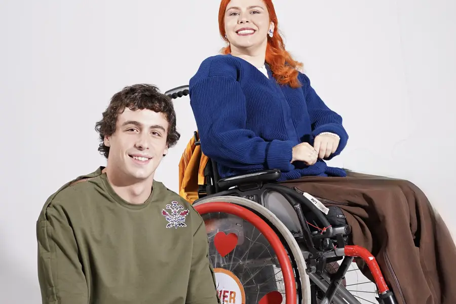 A smiling woman with red hair sits in a decorated wheelchair beside a smiling man in an olive sweatshirt, both posing against a plain light background