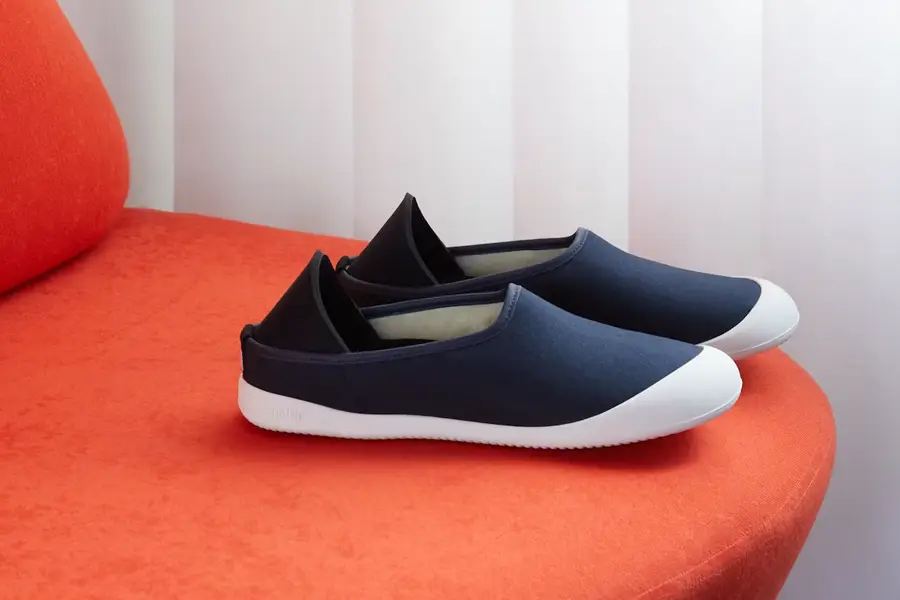 A pair of navy blue slip-on sneakers with white rubber soles and black heel panels, placed on a curved orange velvet ottoman against a white curtained background.