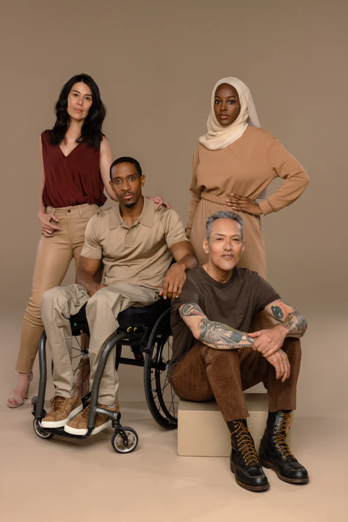 Vertical group portrait of four diverse adults modeling stylish earth-tone adaptive outfits, featuring a seated Black man in a wheelchair, a tattooed man on a block, and two women standing against a neutral brown studio background.