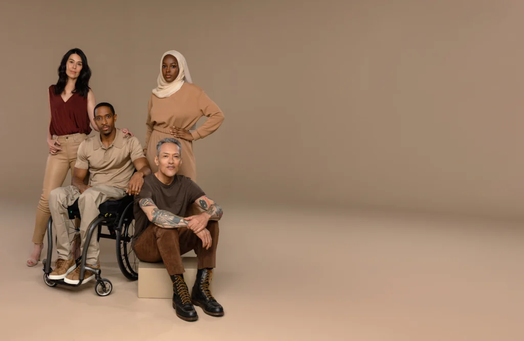 Wide banner layout featuring a diverse group modeling inclusive, earth-toned Adaptt Apparel clothing, including a Black man in a wheelchair and a woman in a hijab, against a solid beige background with ample copy space.