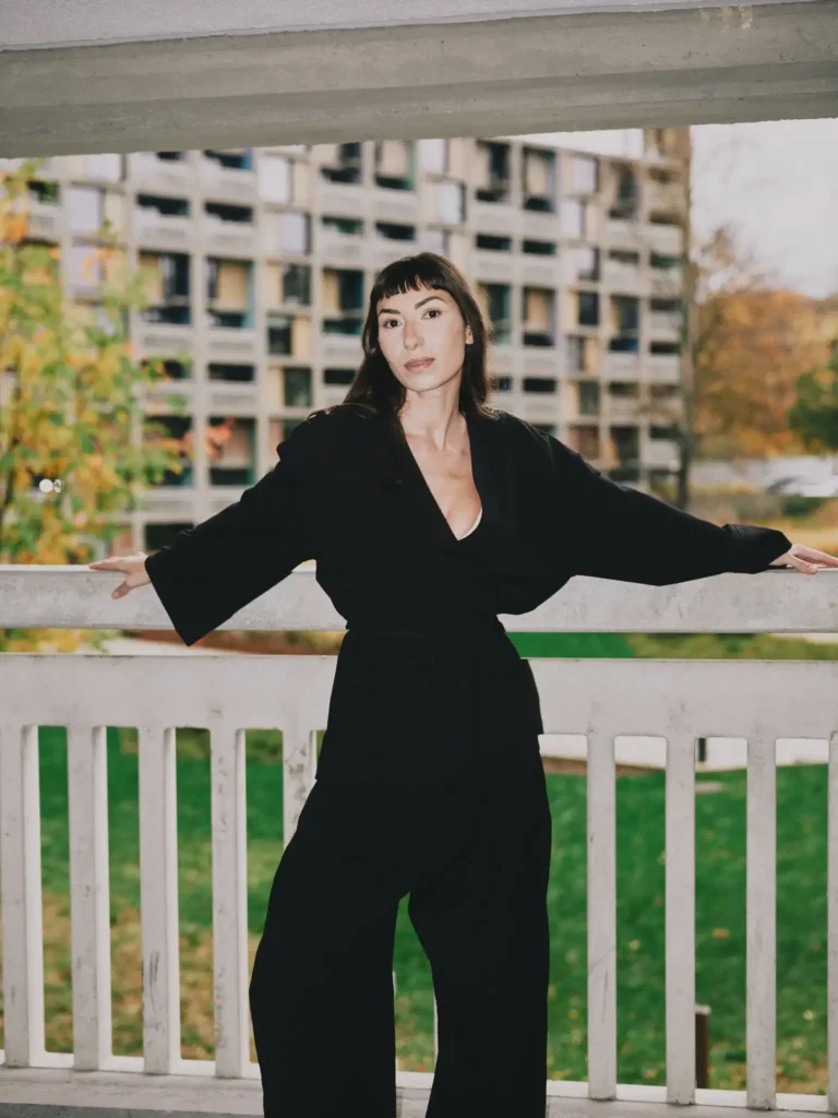 A woman wearing an oversized black linen wrap shirt and wide-leg trousers leaning against a white concrete balcony railing, with a brutalist apartment building background.