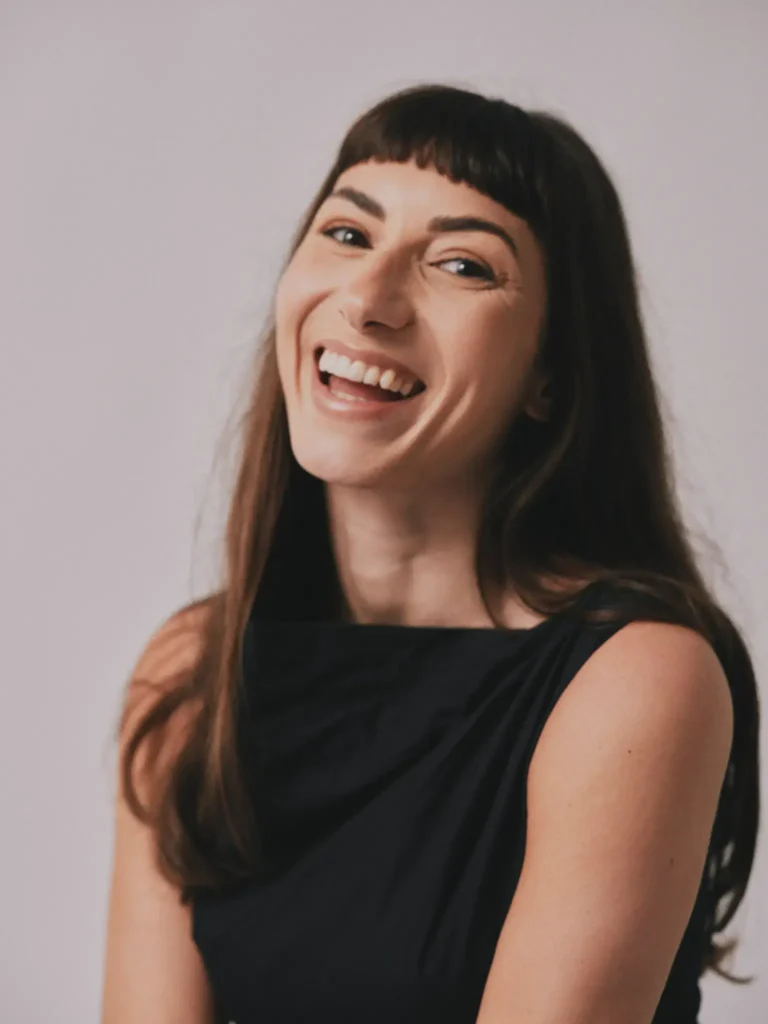 Close-up portrait of a woman with straight bangs and long brown hair laughing candidly, wearing a black sleeveless top against a simple off-white background.