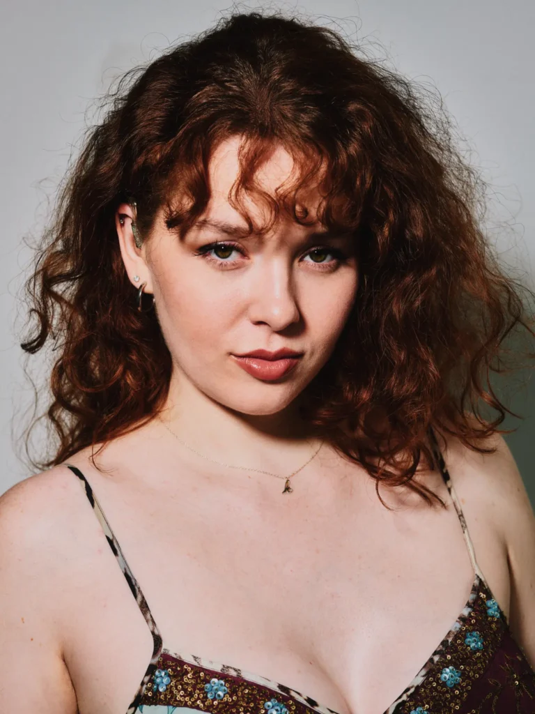 Studio headshot of a woman with curly auburn hair, dramatic lip liner, and visible silver cartilage earrings, wearing a sequined animal print strap top against a grey background.