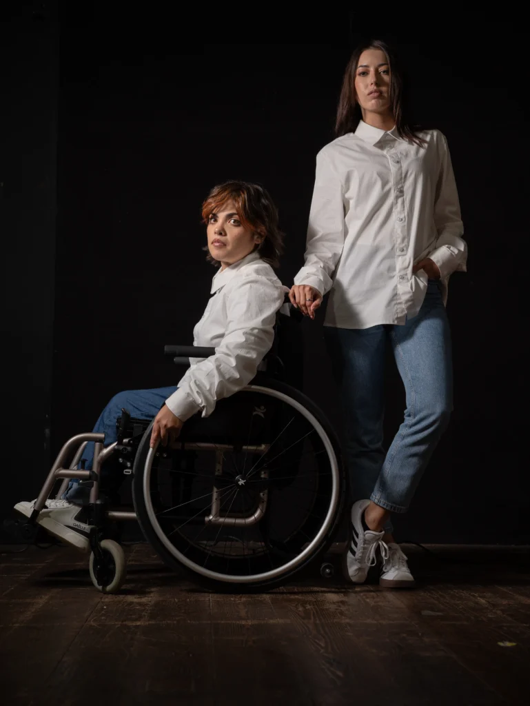 Studio fashion photograph of two female models against a black backdrop, one sitting in a wheelchair and the other standing beside her, both wearing classic white dress shirts and blue jeans.
